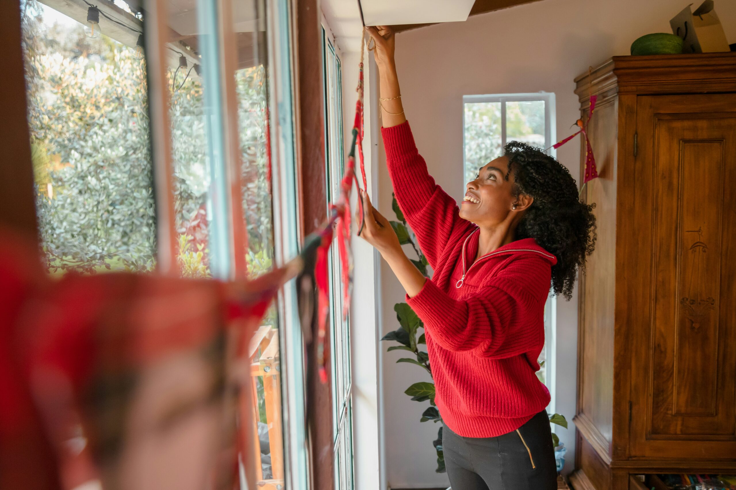 Kvinna som sätter upp juldekorationer inför julafton. Röda detaljer, vid fönstret.